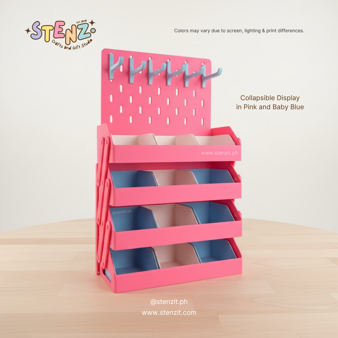 Pink and baby blue collapsible display stand with multiple tiered compartments and a pegboard back panel with hooks, shown on a light wood surface against a plain light background. Text on the image notes the product is collapsible in pink and baby blue.