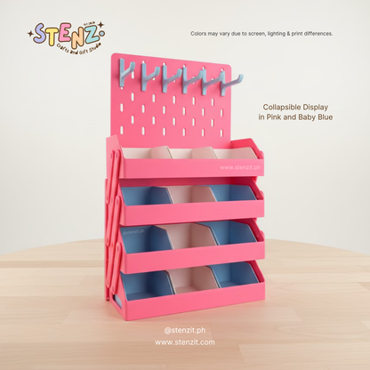 Pink and baby blue collapsible display stand with multiple tiered compartments and a pegboard back panel with hooks, shown on a light wood surface against a plain light background. Text on the image notes the product is collapsible in pink and baby blue.